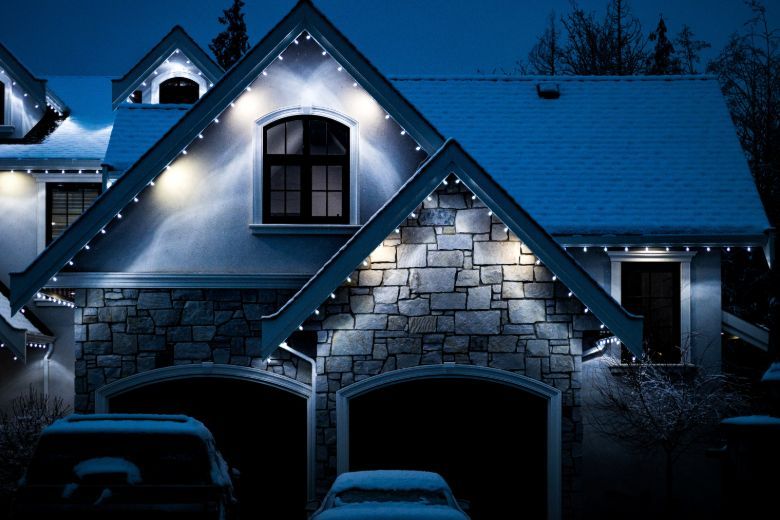 Snow-covered house at night with glowing Christmas lights outlining the roof. Stone facade and dark garage doors add contrast to the winter scene.