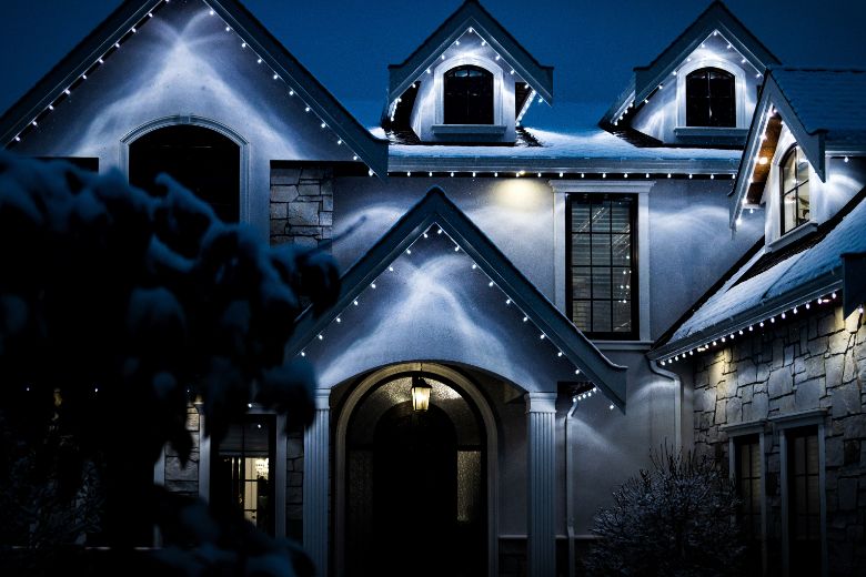 A large house with white Christmas lights outlining the roofline and gables. Snow dusts the roof and surrounding shrubbery, creating a serene winter night scene.