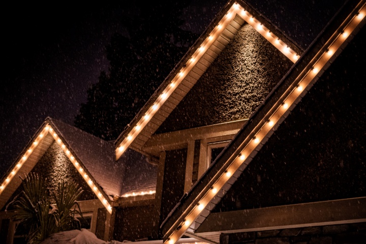 A snow-dusted home glows with warm Christmas lights outlining its roofline against the dark night sky. Gentle snowflakes fall, adding a magical touch to this cozy winter scene.